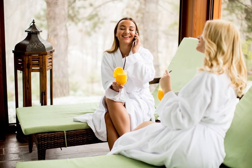 Two cheerful women in bathrobes drinking juice in spa center